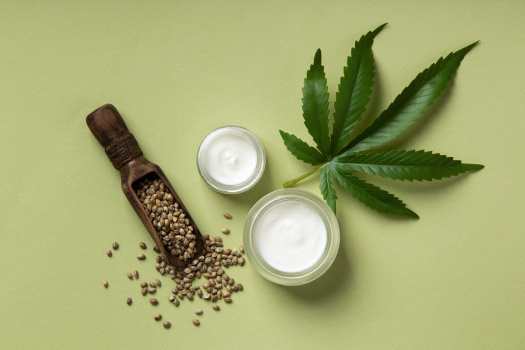 A top-down view of a CBD oil bottle and cosmetic jars with fresh hemp leaves on a grey surface, perfect for a shopify CBD store display.