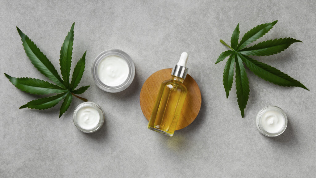 An overhead view of a CBD oil dropper and jars of white topical cream with hemp leaves, highlighting a diverse WooCommerce CBD product line.